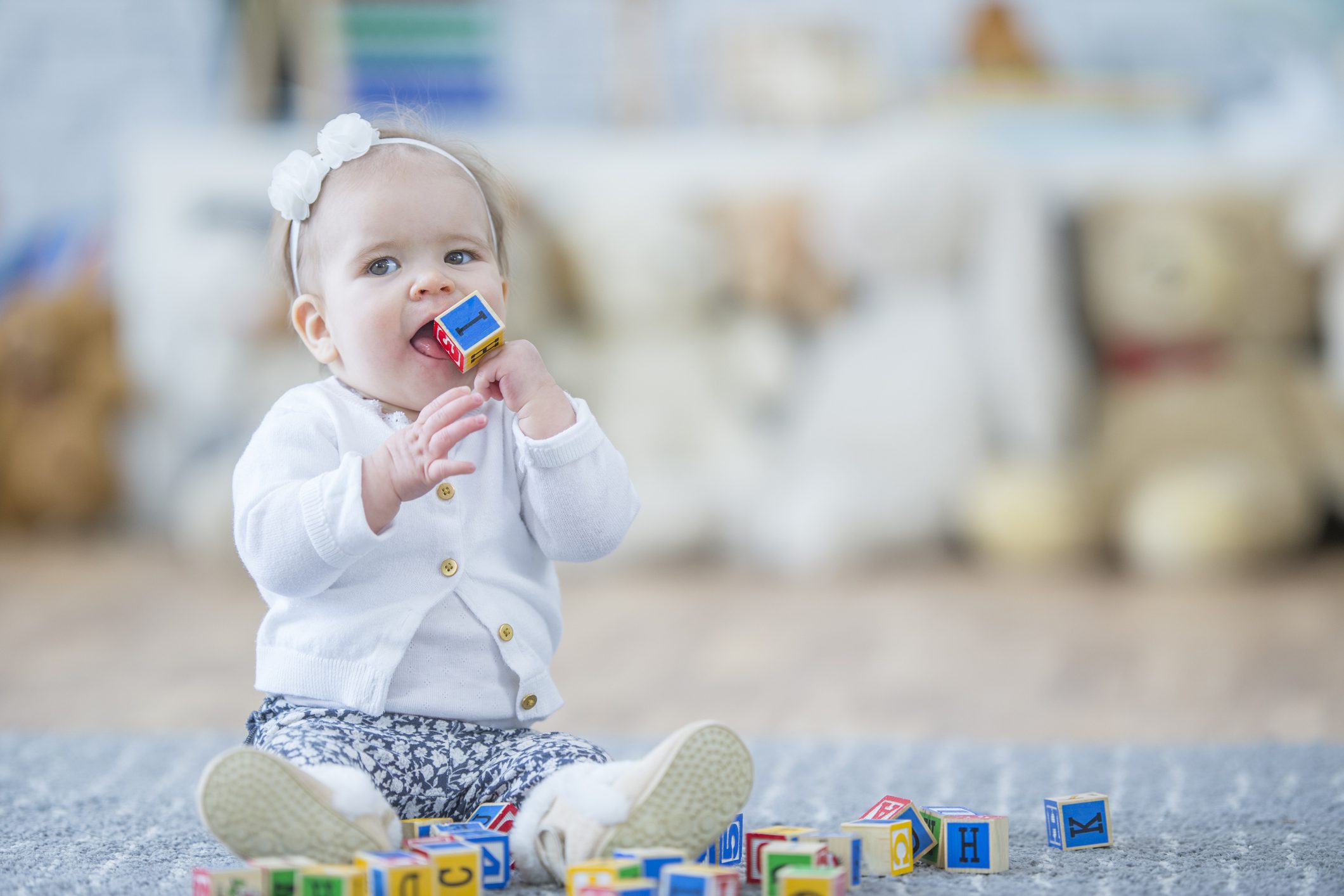 A baby girl is indoors in a daycare center. She is playing with toy blocks and trying to put one into her mouth.